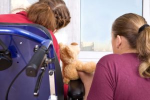 Back of girl in wheelchair holding a stuffed bear and sitting with woman 