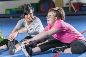 A fitness instructor in a gym working with a teenage girl with down syndrome. They are sitting on the floor, stretching.