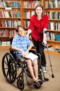 a boy student in a wheelchair next to a girl student standing on crutches in a library