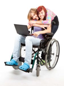 a young lady hugging another sitting in a wheelchair holding a laptop