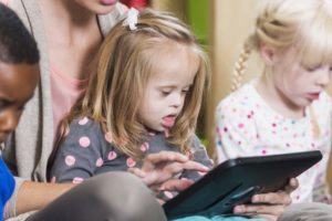 A little white girl with down syndrome, sitting on the lap of a woman, her mother or teacher, looking at a digital tablet. The woman is cropped and unrecognizable. Two other children are sitting beside them.