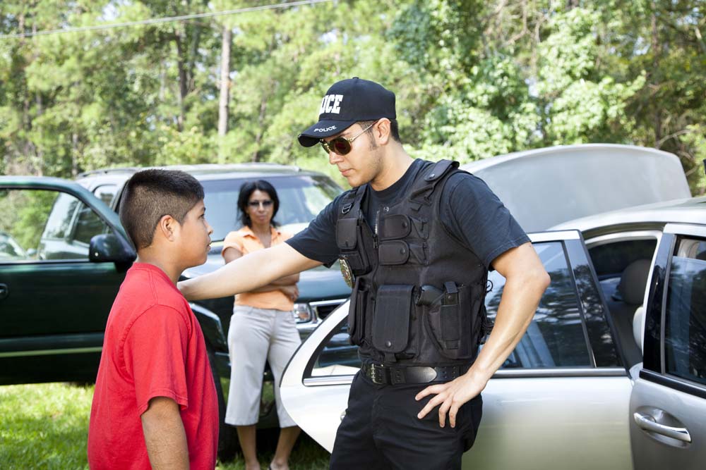 A Latino policeman questions an elementary aged Latino boy, his hand on his shoulder, while the Mom watches in the background.