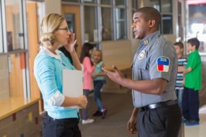 Black/African-American police officer talks seriously to concerned white school teacher with children in background.