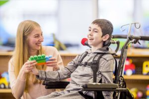 A smiling teacher helps an elementary school white boy with a physical disability play with plastic block toys.