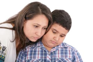 A Latino Mother comforts her adolescent son. Both look down with sad expressions.